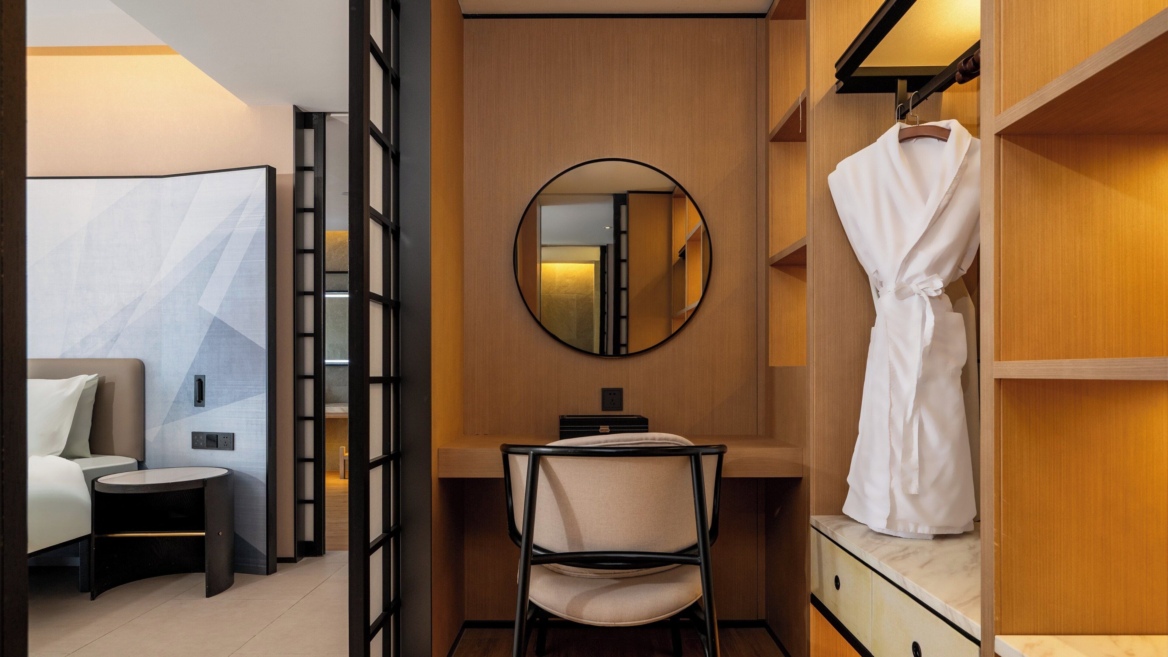A hotel room with a bed, a desk, a chair, a robe hanging on a rack, and a mirror on the wall.