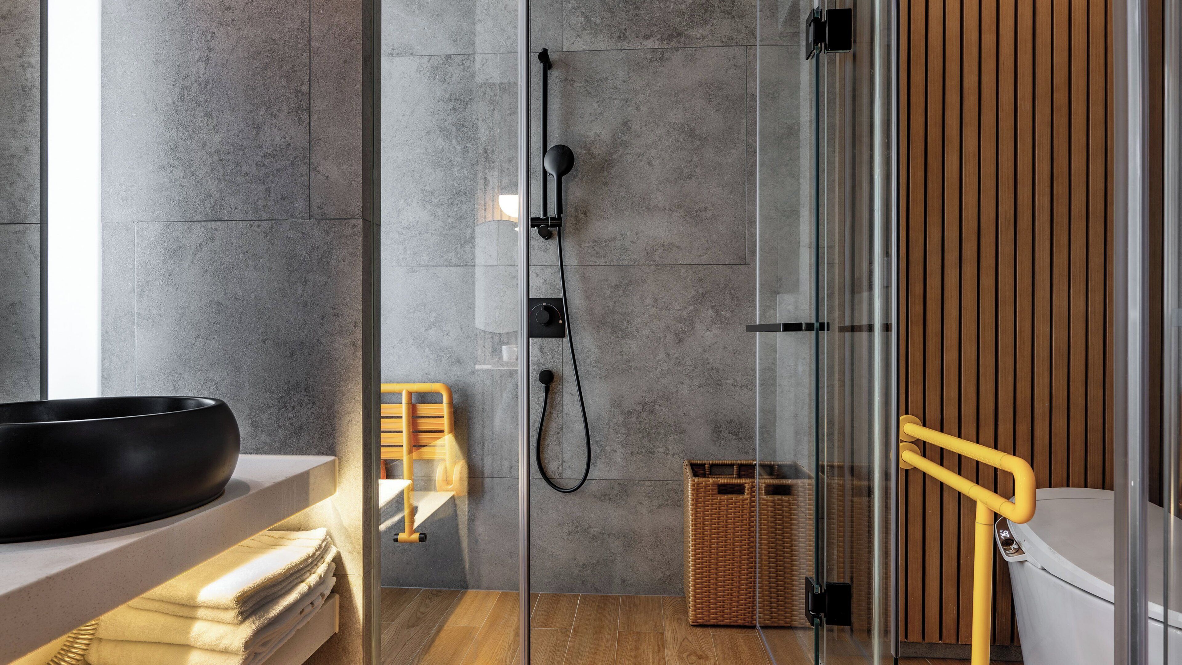 A modern bathroom with a glass shower enclosure, a wooden chair, a black basin, and a wicker basket.