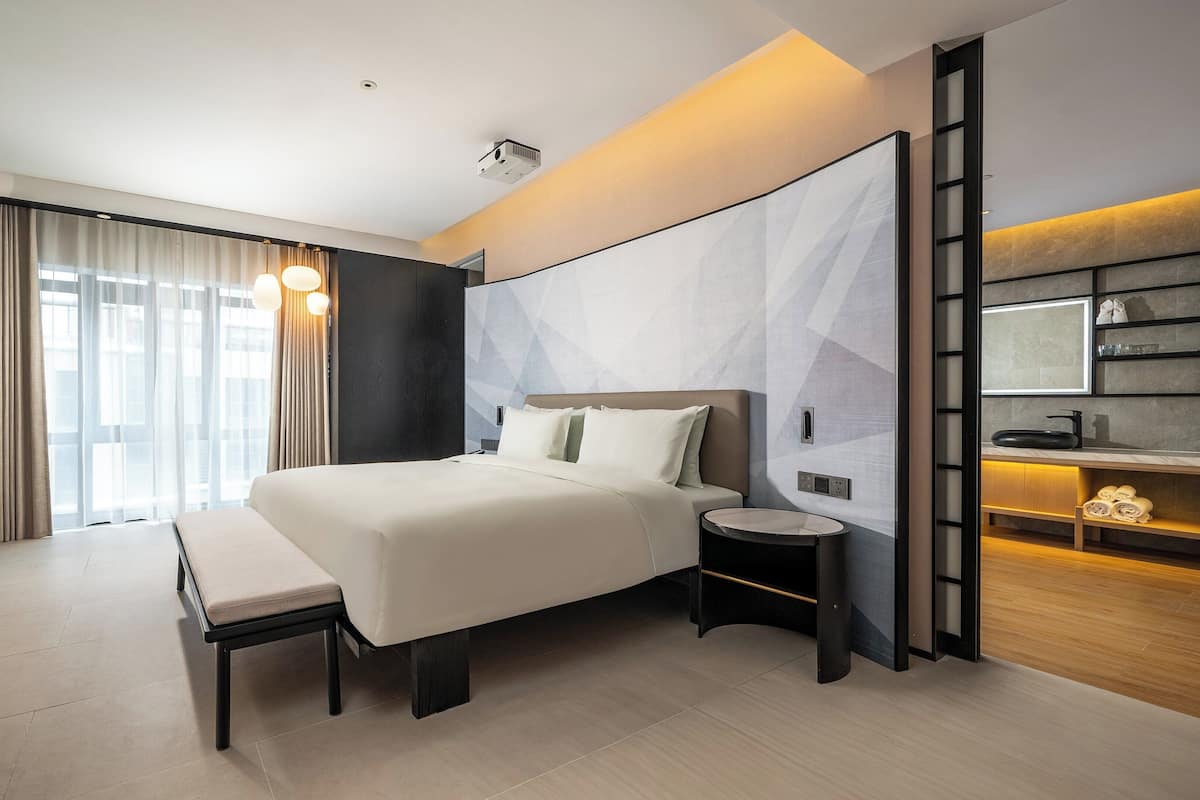 Ein modernes Hotelzimmer mit einem großen Bett, einem Nachttisch, einer Bank und einem integrierten Schreibtisch mit Spiegel und Regalen.