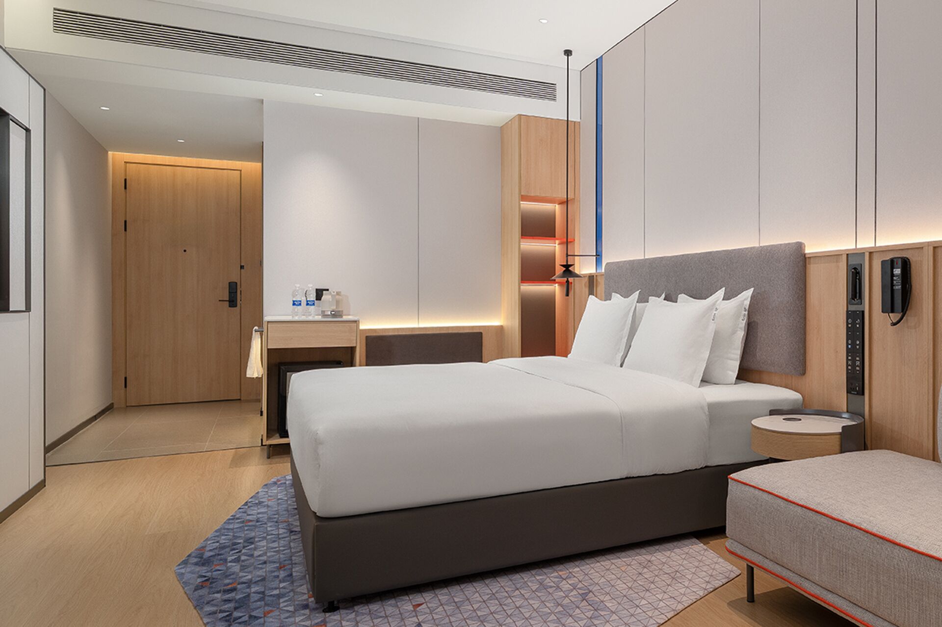 A modern hotel room with a large bed, bedside table, and a desk with a chair. There is a wardrobe, a mirror, and a phone on the wall.