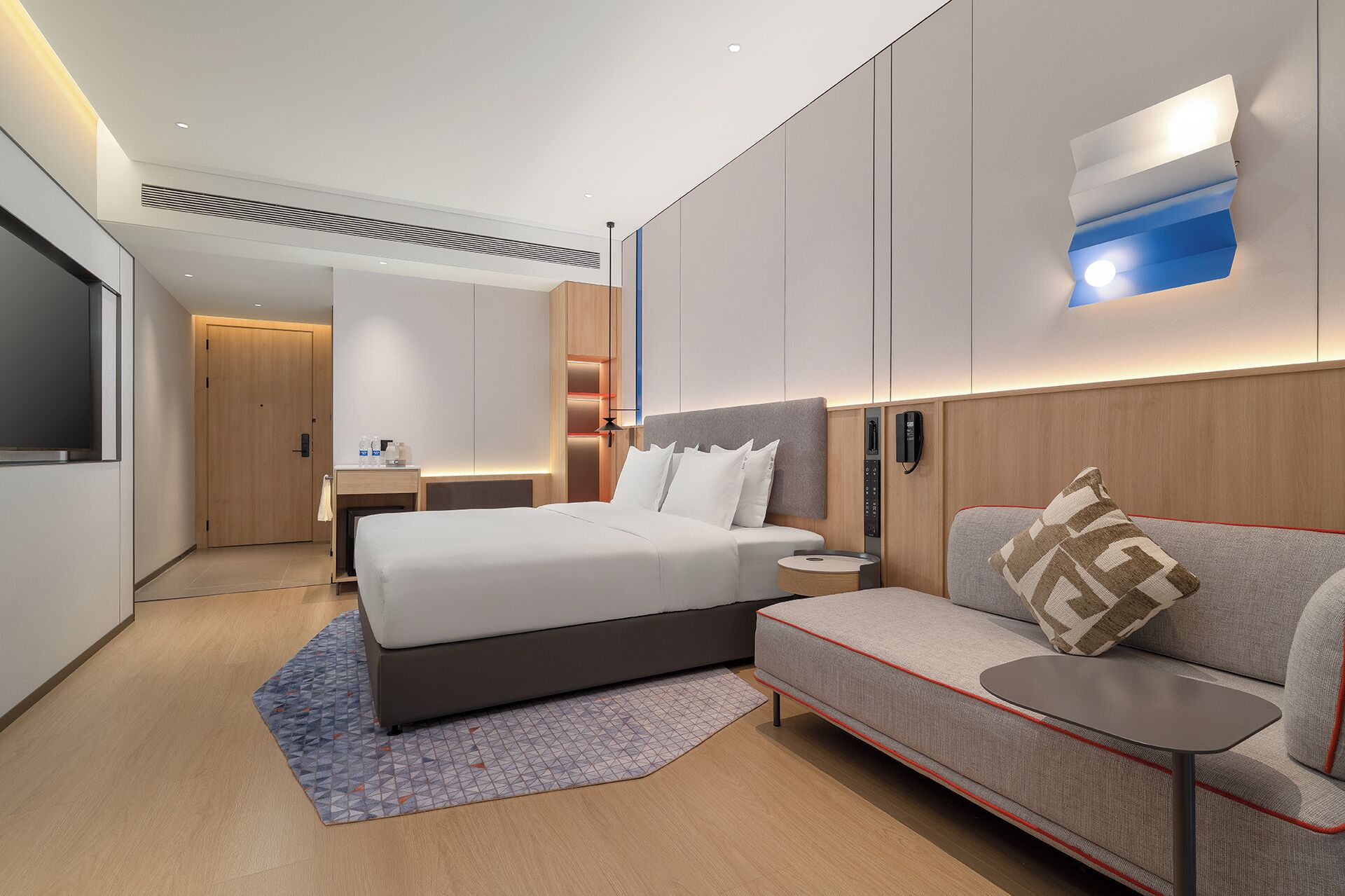 A modern hotel room with a large bed, a sofa, a small table, and a wall-mounted light fixture.
