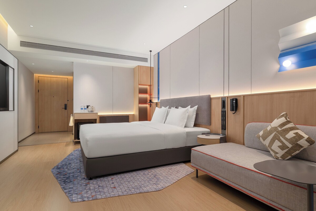 A modern hotel room with a large bed, a sofa, a desk, and a flat-screen TV.