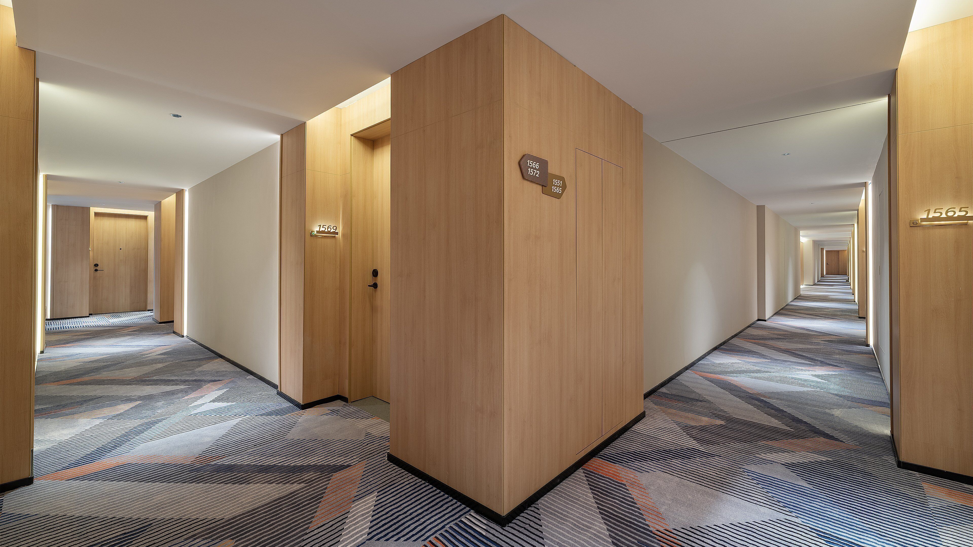Een hotelgangen met houten deuren, genummerde kamerdeuren en een kamerbreed tapijt met patroon.