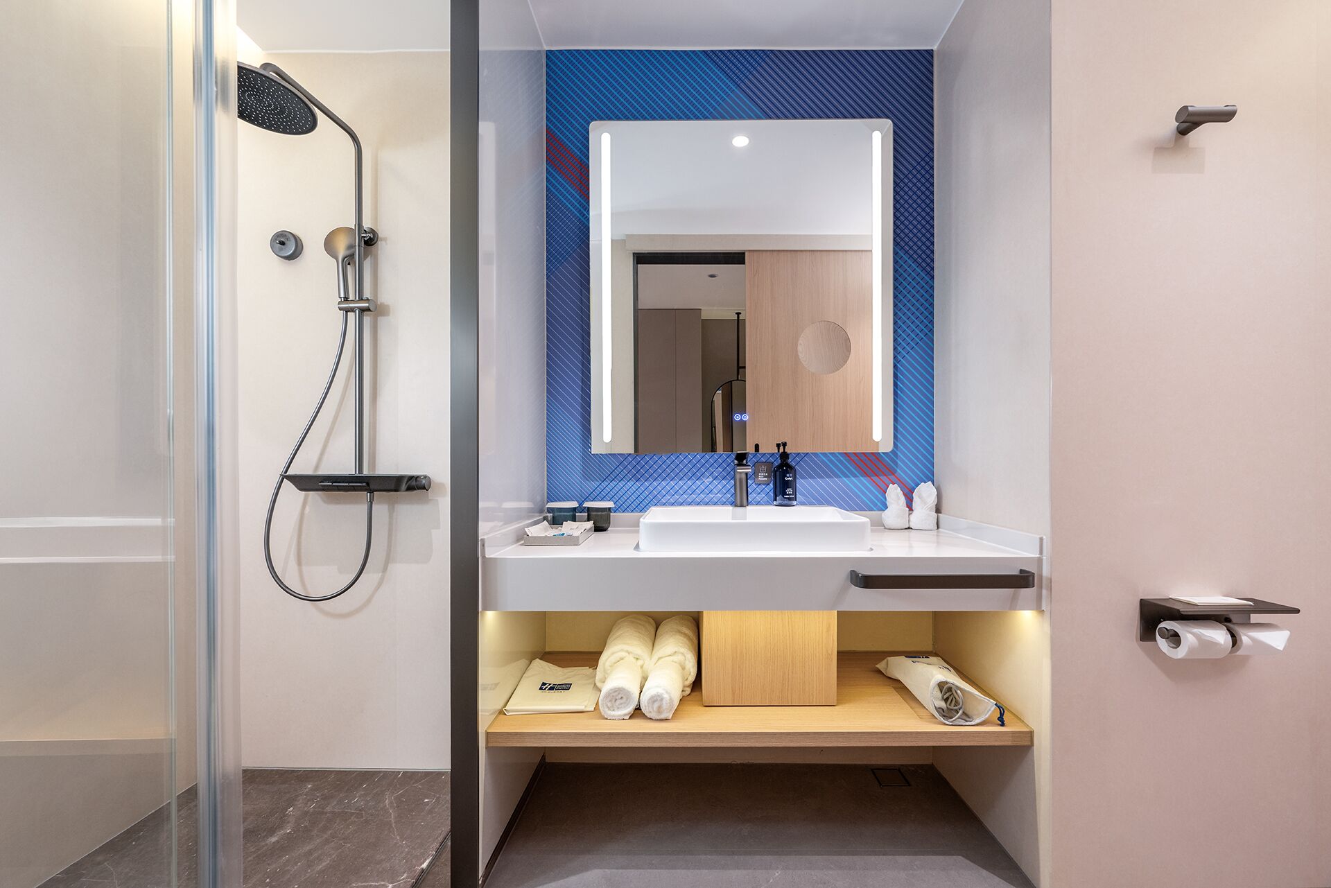 Een moderne badkamer met een douchecabine van glas, een witte wastafel met een spiegel erboven en een houten plank met handdoeken en toiletartikelen.
