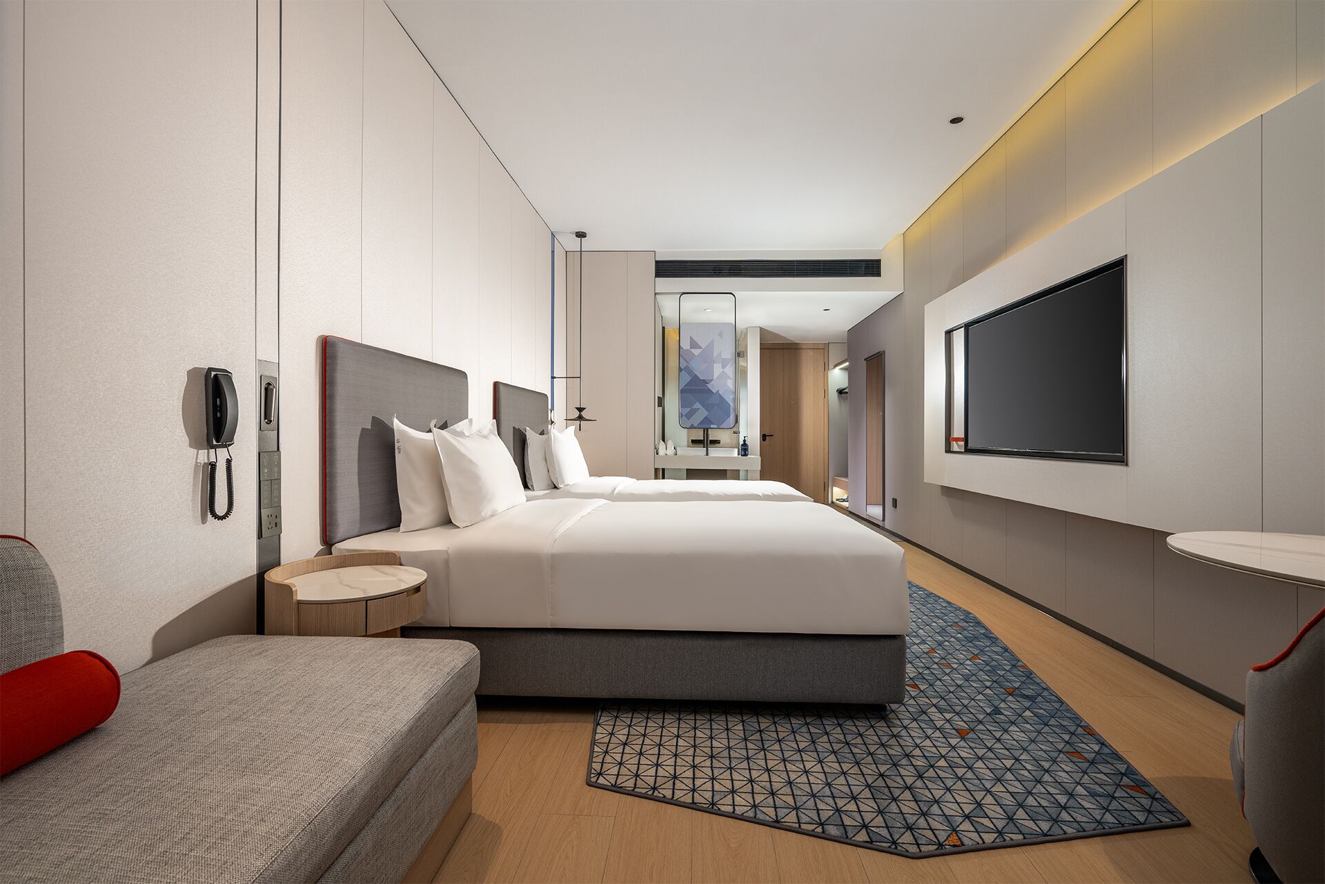 A modern hotel room with a large bed, bedside tables, a desk, a TV, and a phone on the wall.