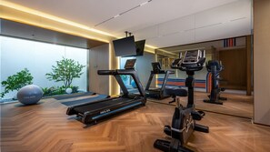 A modern gym with a treadmill, elliptical, and stationary bike, wooden flooring, and a large mirror reflecting the room.