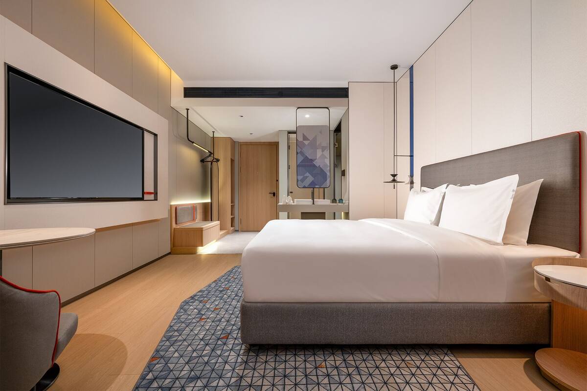 A modern hotel room with a large bed, a flat-screen TV, a desk, and a bathroom with a mirror and sink.