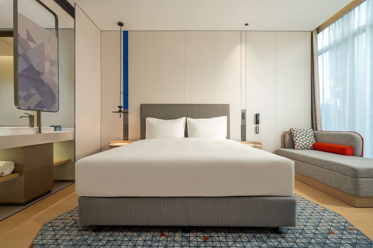 A modern hotel room with a large bed, a bedside table, a nightstand, a sofa, a patterned rug, and a large window with curtains.
