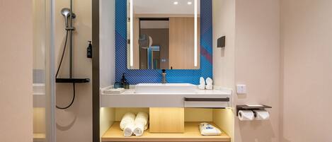 Un baño moderno con ducha de vidrio, un espejo grande, lavamanos con tocador de madera y un dispensador de papel higiénico.