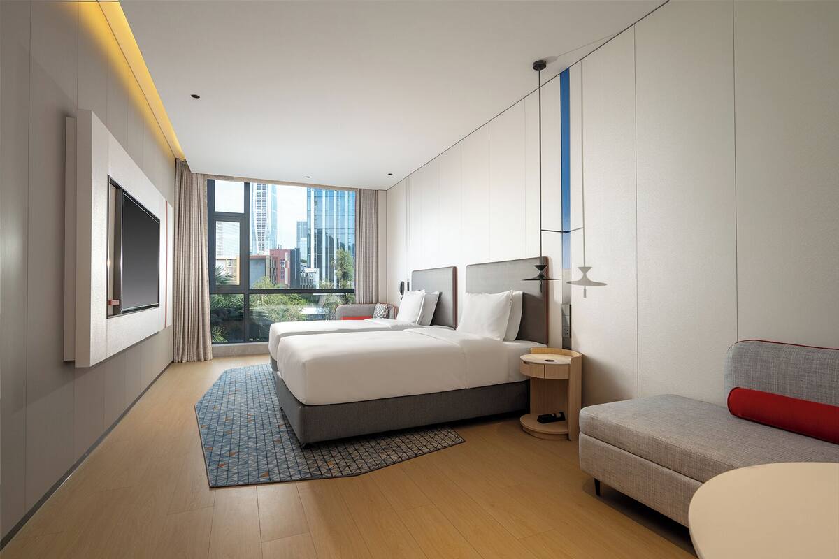 A modern hotel room with a large bed, a sofa, a small table, and a view of the cityscape.