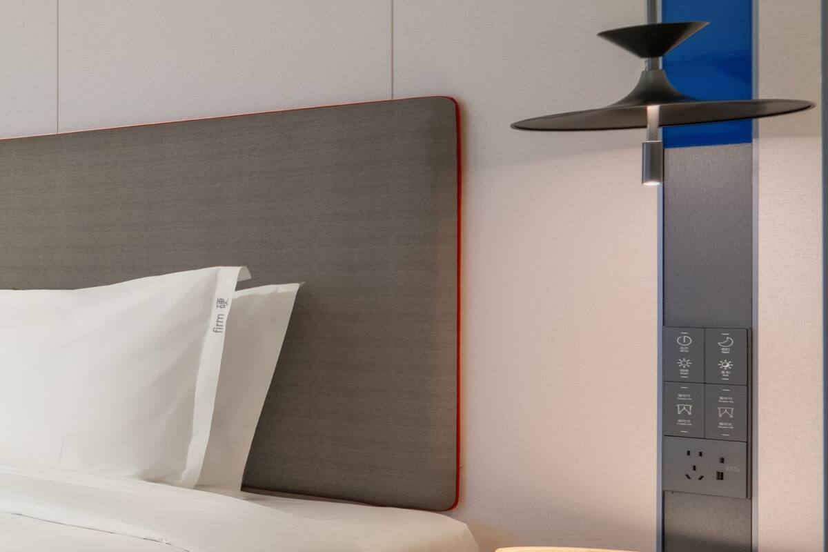 A modern hotel room with a grey headboard, white bedding, and a control panel with Japanese text.