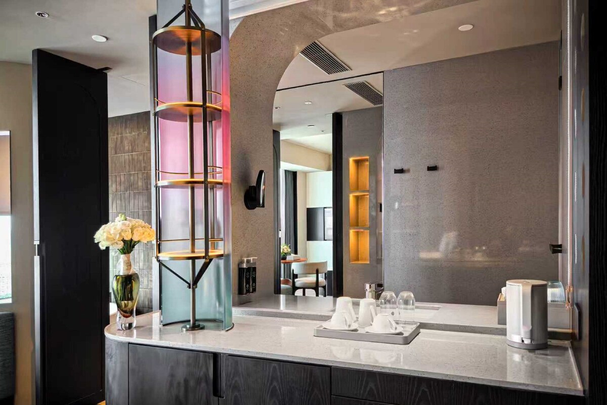 A modern bathroom with a large mirror, a round vanity with a dark wood finish, and a unique cylindrical light fixture.