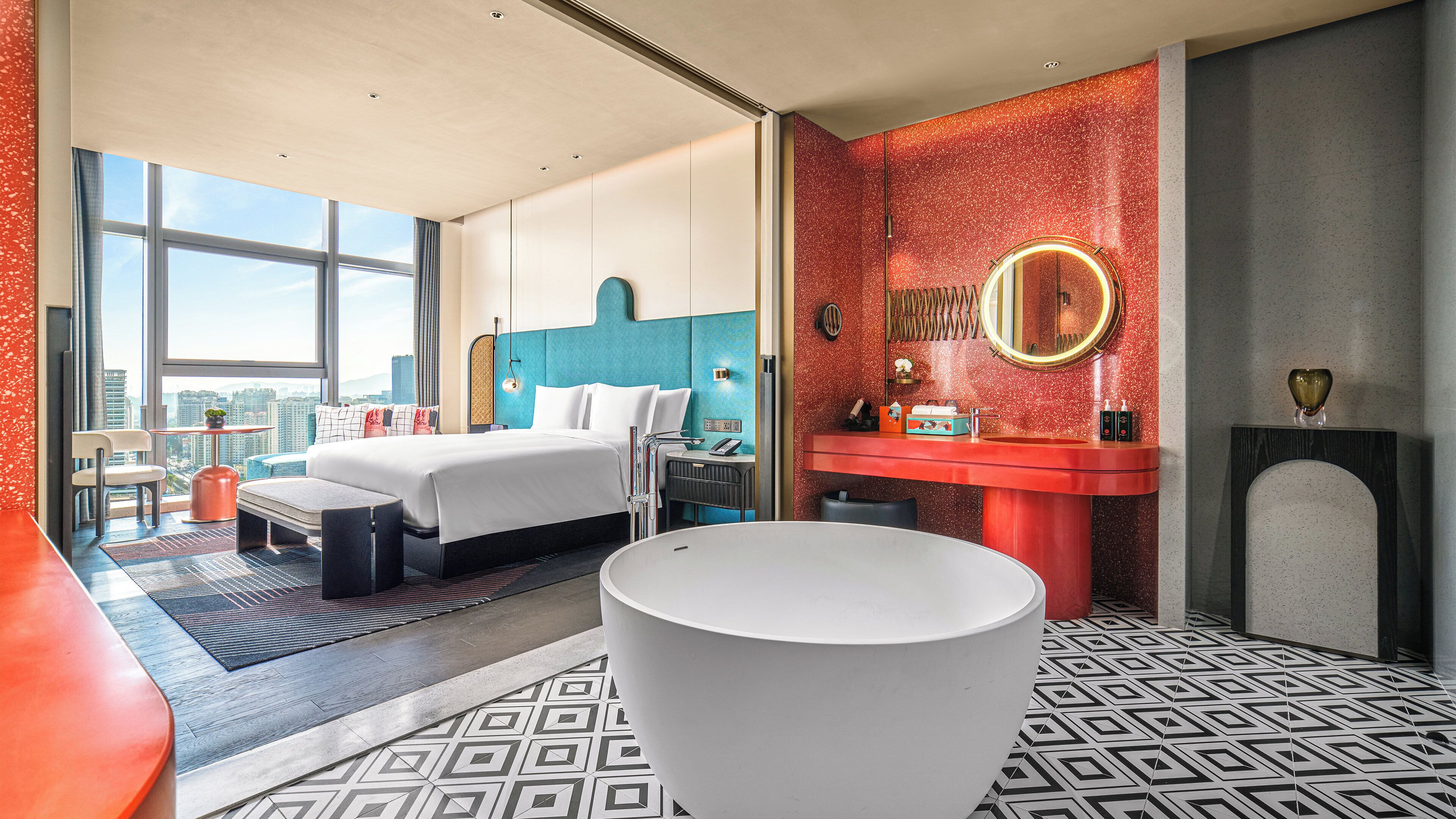 A modern hotel room with a large bed, a sitting area, a bathroom with a vanity, and a large window offering a city view.