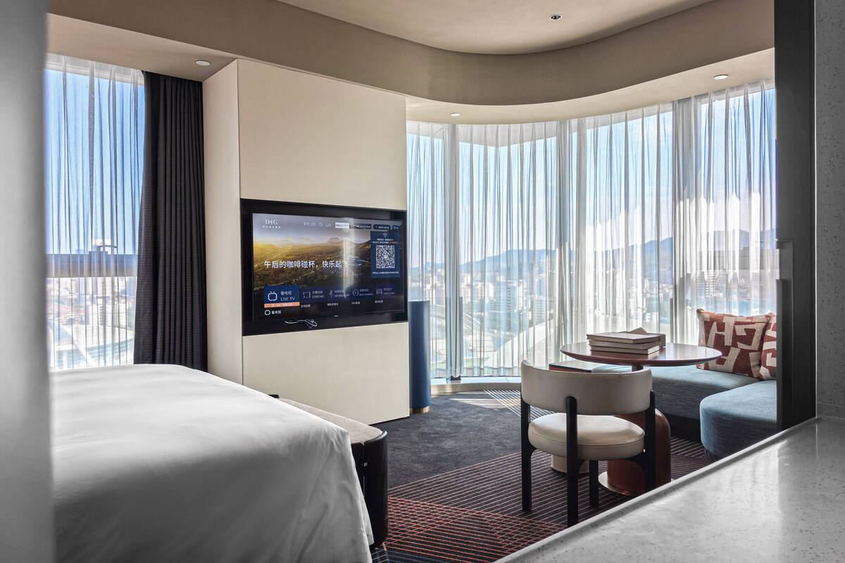 A hotel room with a bed, a TV mounted on the wall, a small table with books, and a view of the city through large windows.
