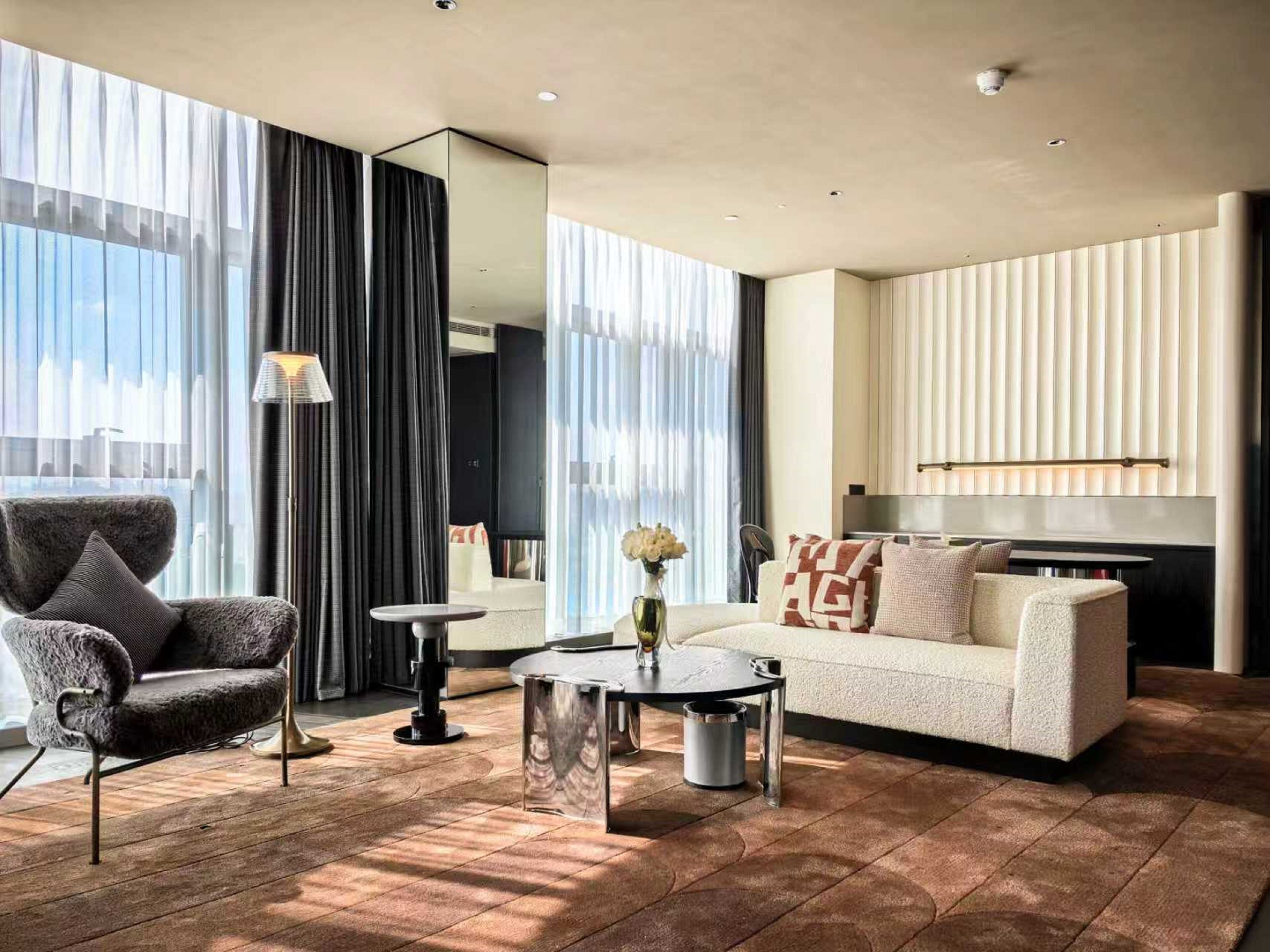 A modern hotel room with a large window, a sofa, a chair, a coffee table, and a mirror.