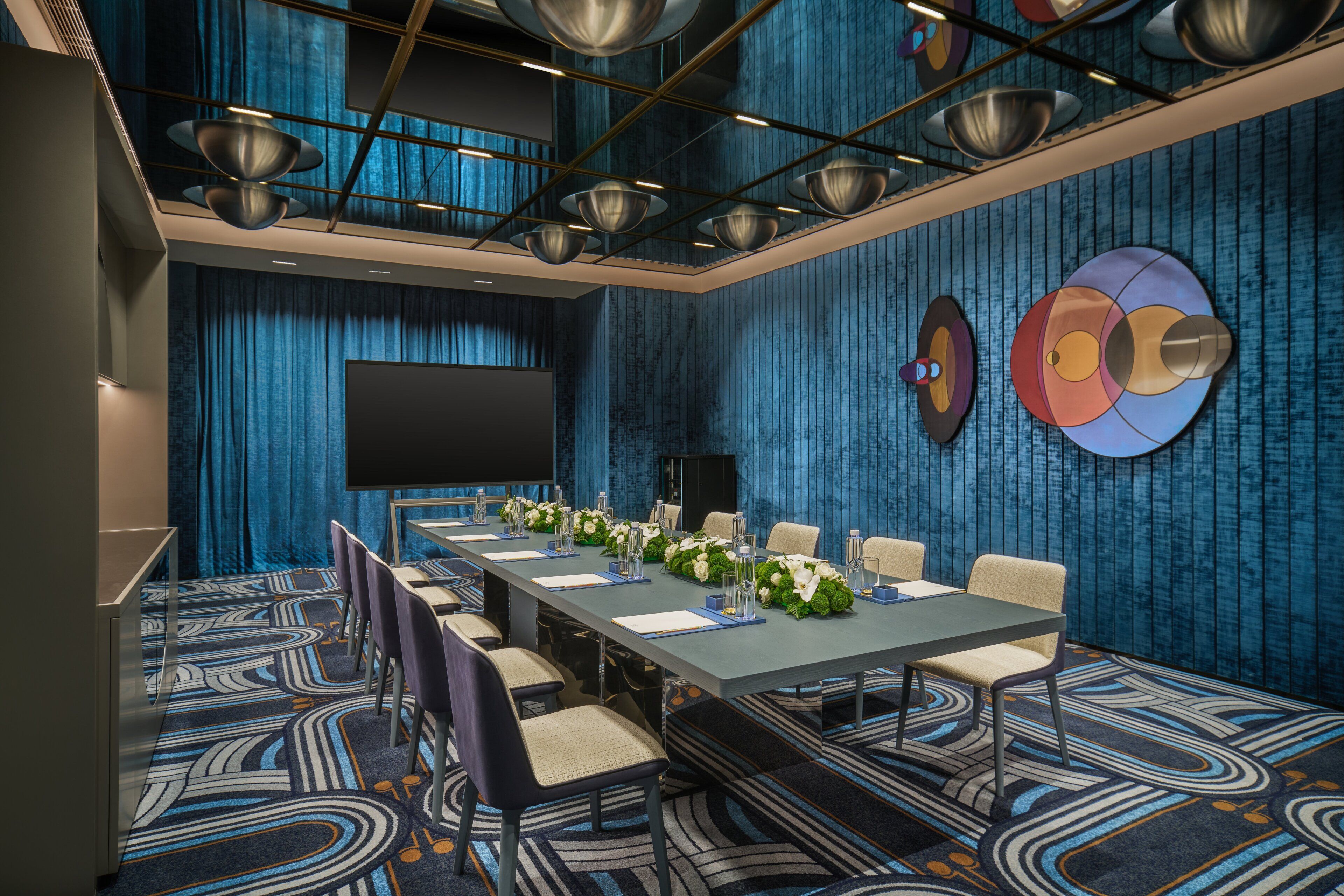 A conference room with a long table, chairs, a projector screen, and a blue patterned carpet.
