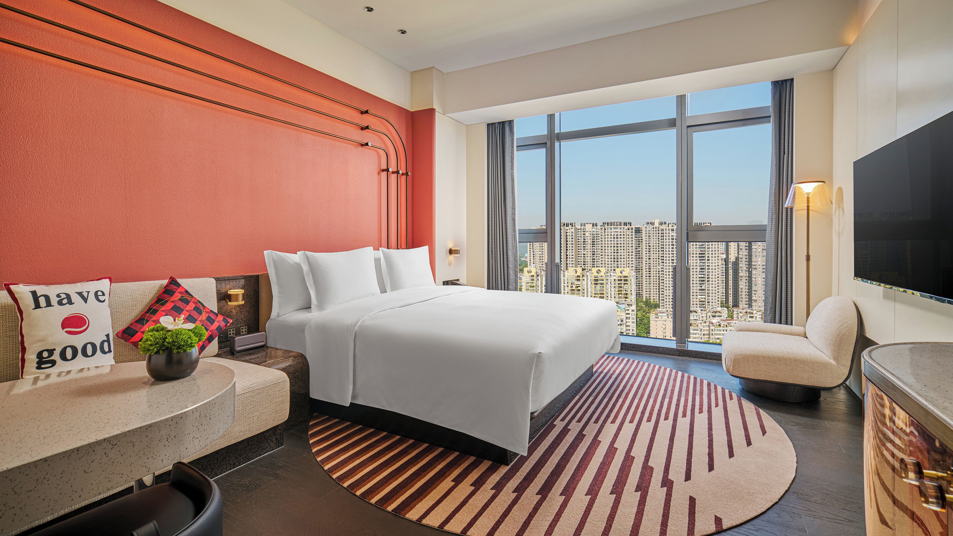 Ein Hotelzimmer mit einem Bett, einer Sitzecke mit Tisch, einem Fernseher und einem großen Fenster mit Blick auf die Stadt.