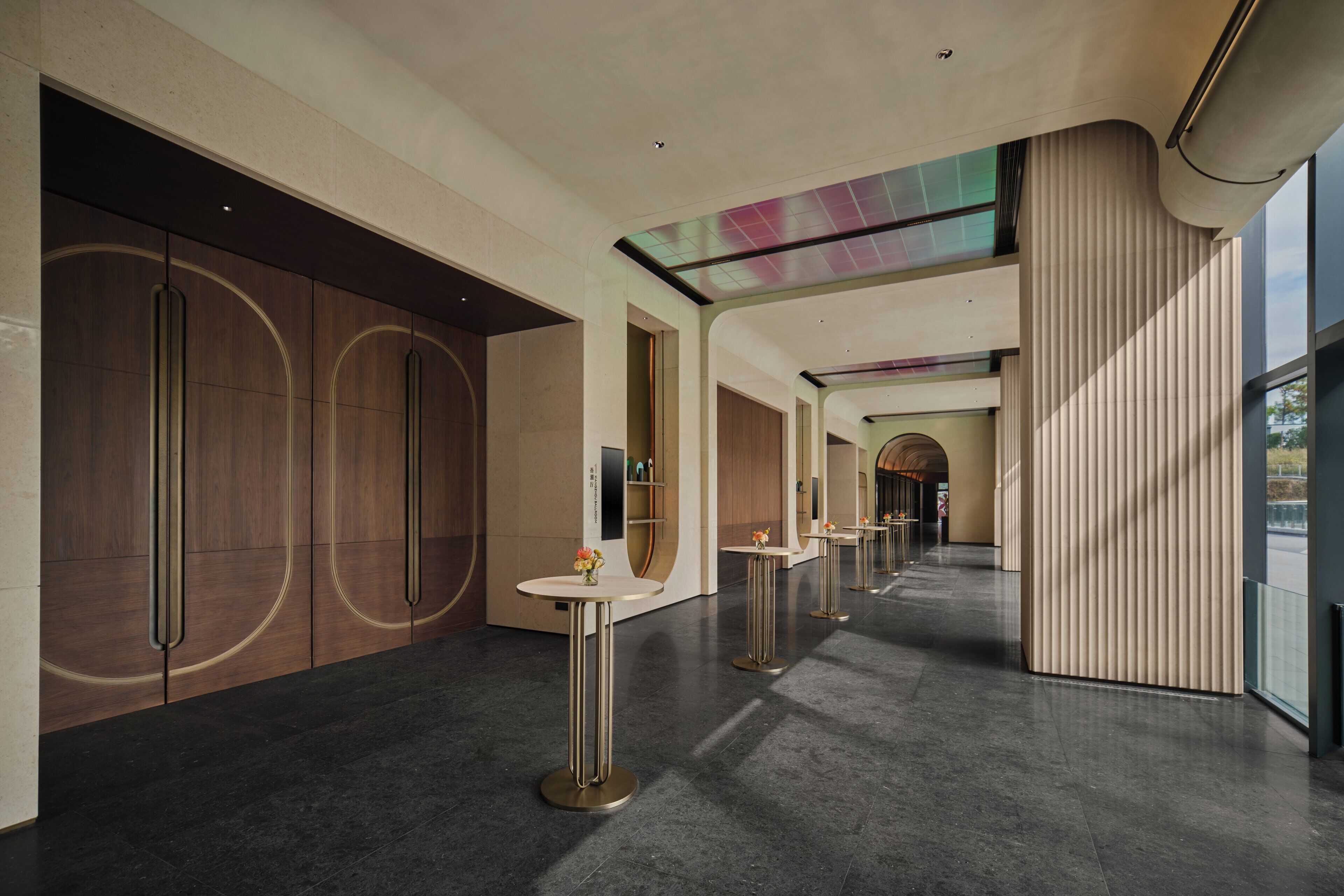A modern hotel corridor with dark tiled flooring, minimalist decor, and a series of round tables with vases.