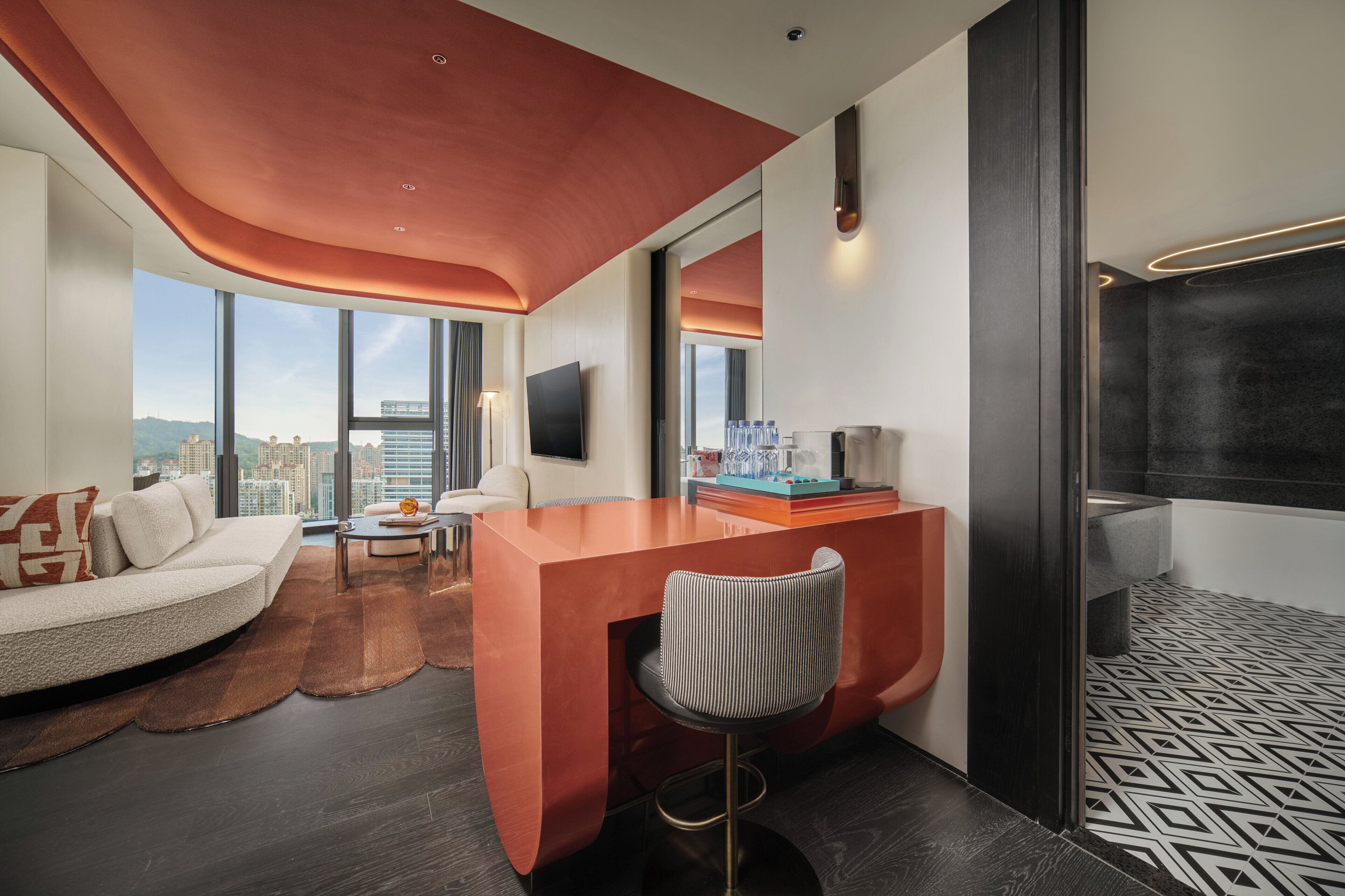 Una habitación de hotel moderna con techo curvo, una zona de bar, un sofá, una silla, un ventanal con vistas a la ciudad y un baño con un diseño geométrico en el suelo.