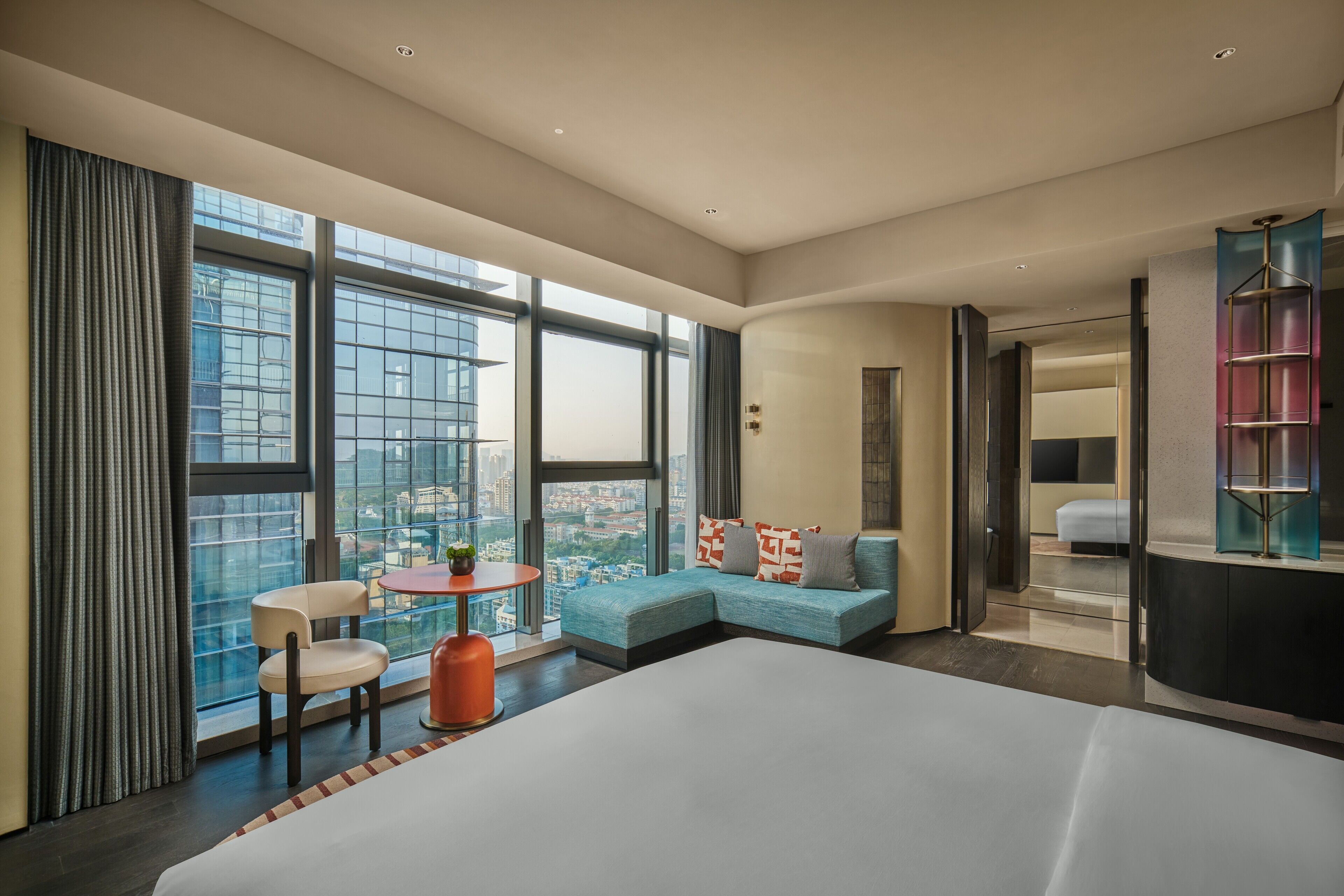 A modern hotel room with a large bed, a sofa, a small table, and a chair. There is a large window offering a city view.