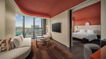 Een moderne hotelkamer met een groot bed, een bank, een bijzettafeltje en een televisie. De kamer heeft uitzicht op de stad door grote ramen.