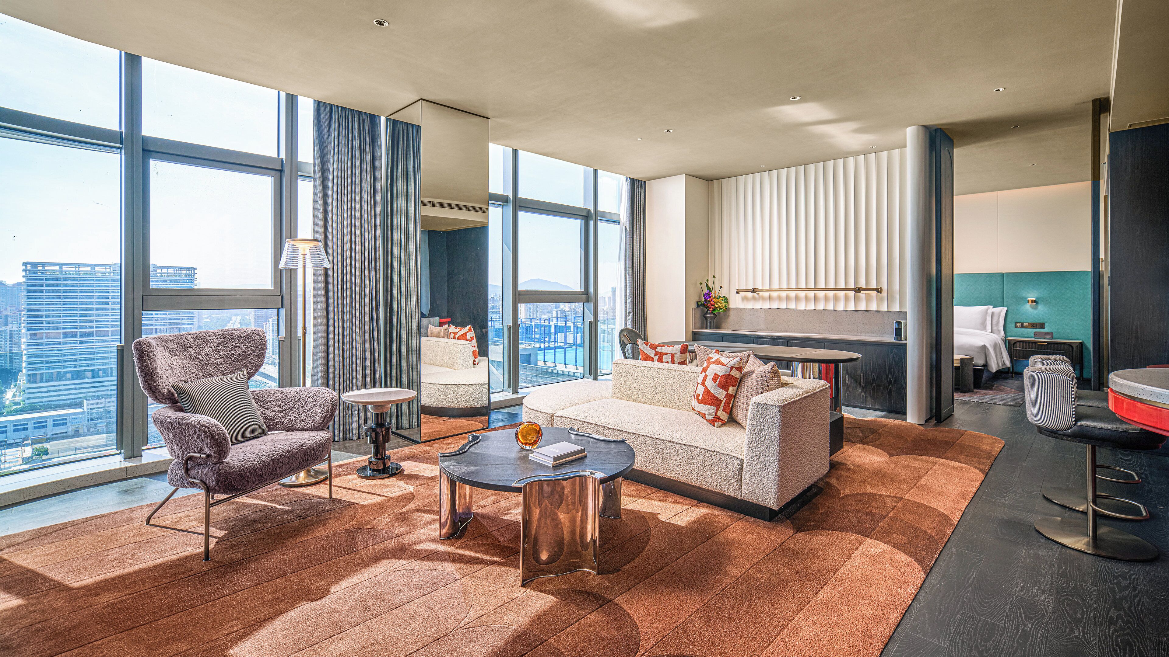 Una habitación de hotel moderna con un ventanal grande, un sofá, una silla, una mesita de centro y una zona de cocina.