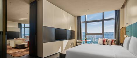 A modern hotel room with a large bed, a flat-screen TV, a seating area with a sofa and armchairs, and a city view through large windows.