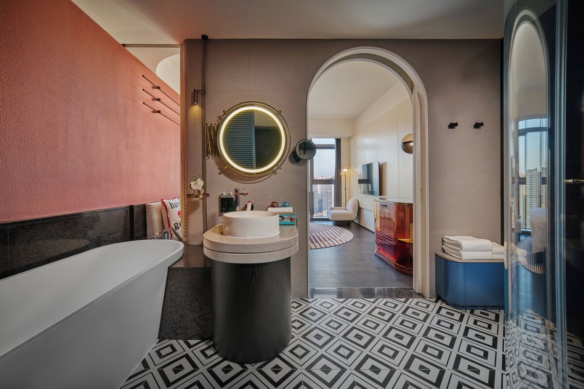 A modern bathroom with a bathtub, a round mirror, and a sink. There is a patterned floor and a view of a cityscape through an archway.