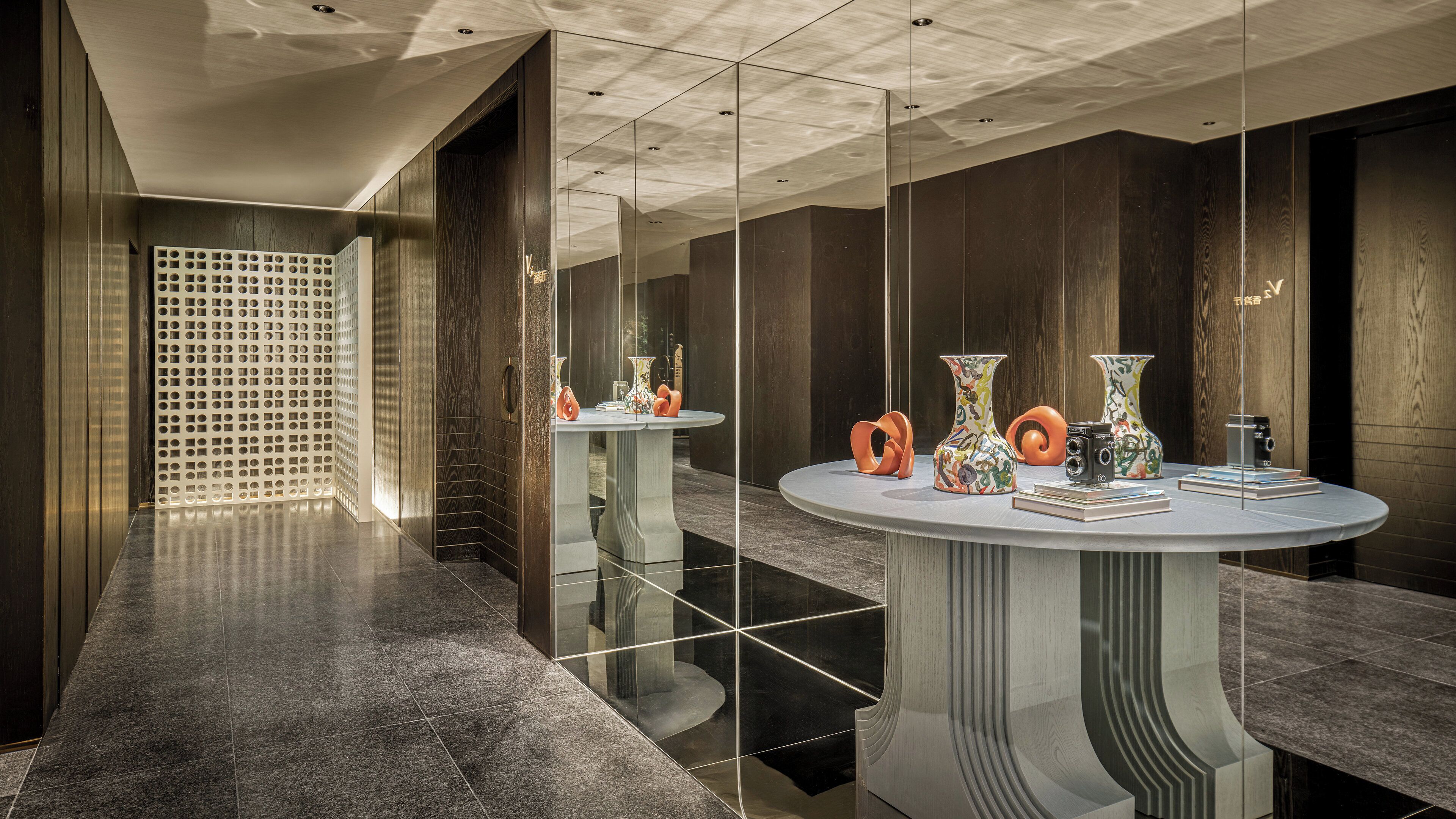 Una hall d'hotel moderna con pavimenti riflettenti, rivestimenti in legno scuro e un tavolo rotondo centrale con oggetti decorativi.