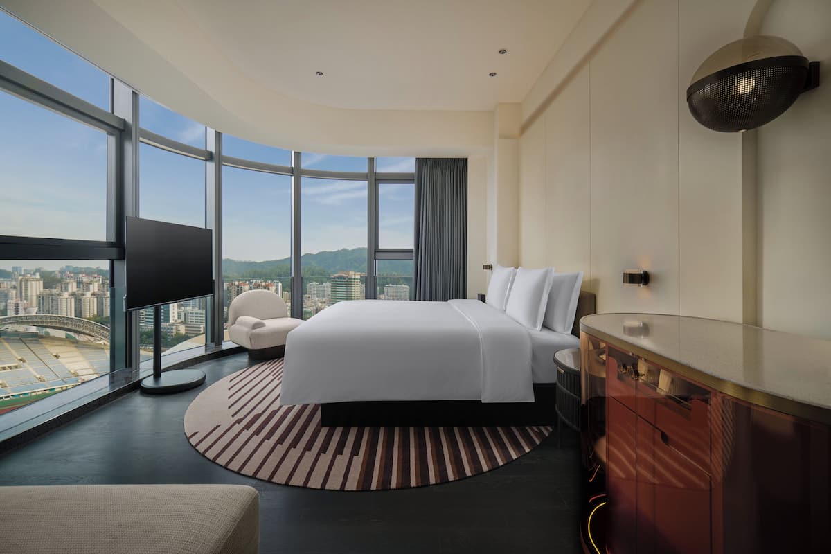 A modern hotel room with a large bed, a TV, a desk, and a view of a cityscape.