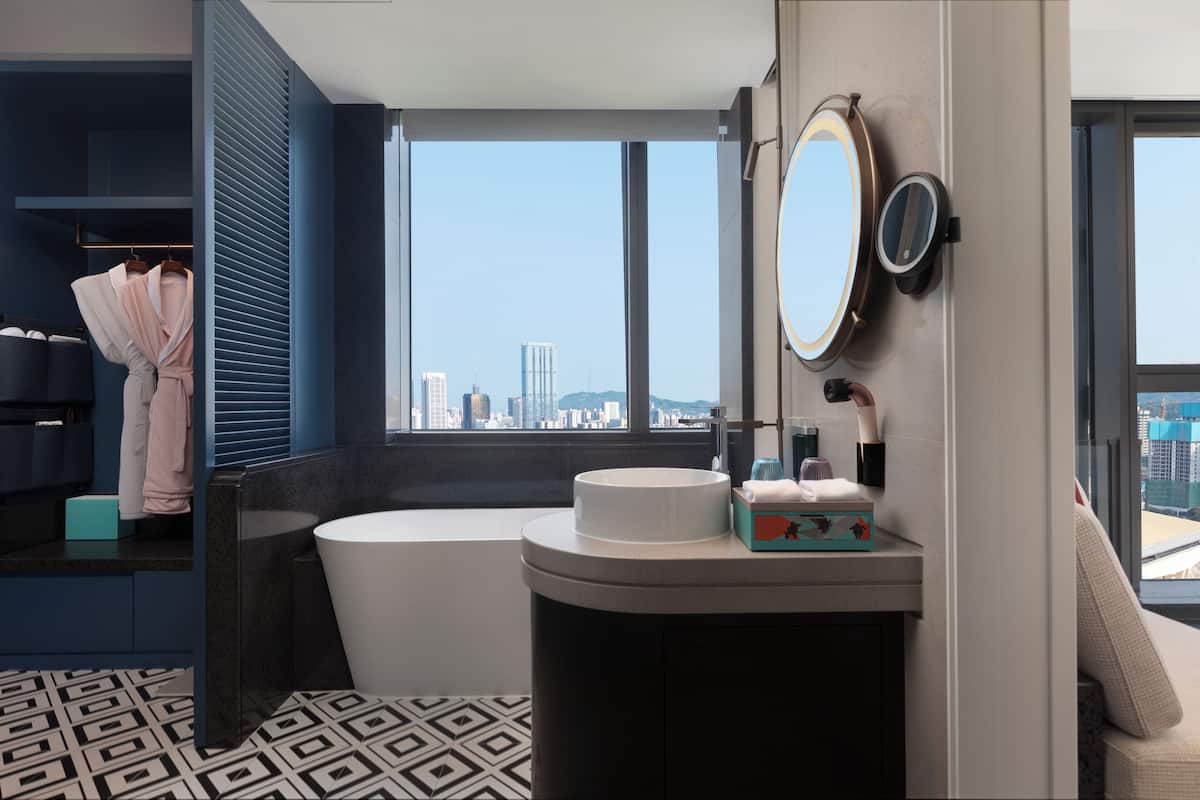 Une salle de bain moderne avec une baignoire, un miroir rond et une fenêtre donnant sur la ville.