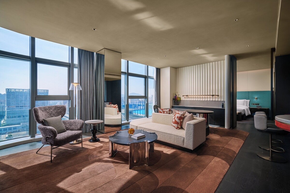 A modern living room with a sofa, armchair, and coffee table, large windows with a city view, and a kitchen area with a bar.