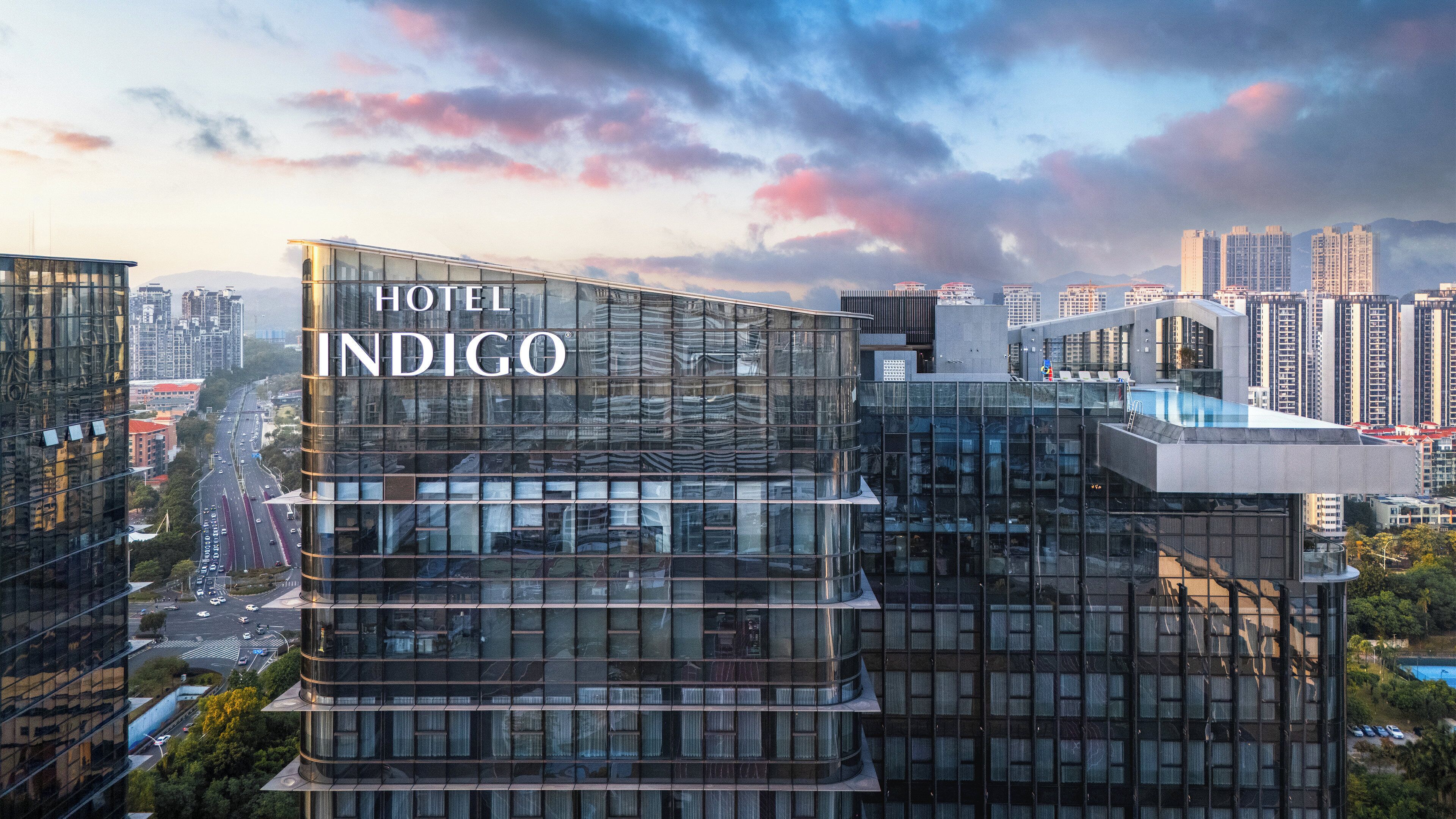 A modern hotel building with a glass facade, labeled 'HOTEL INDIGO,' surrounded by a cityscape.