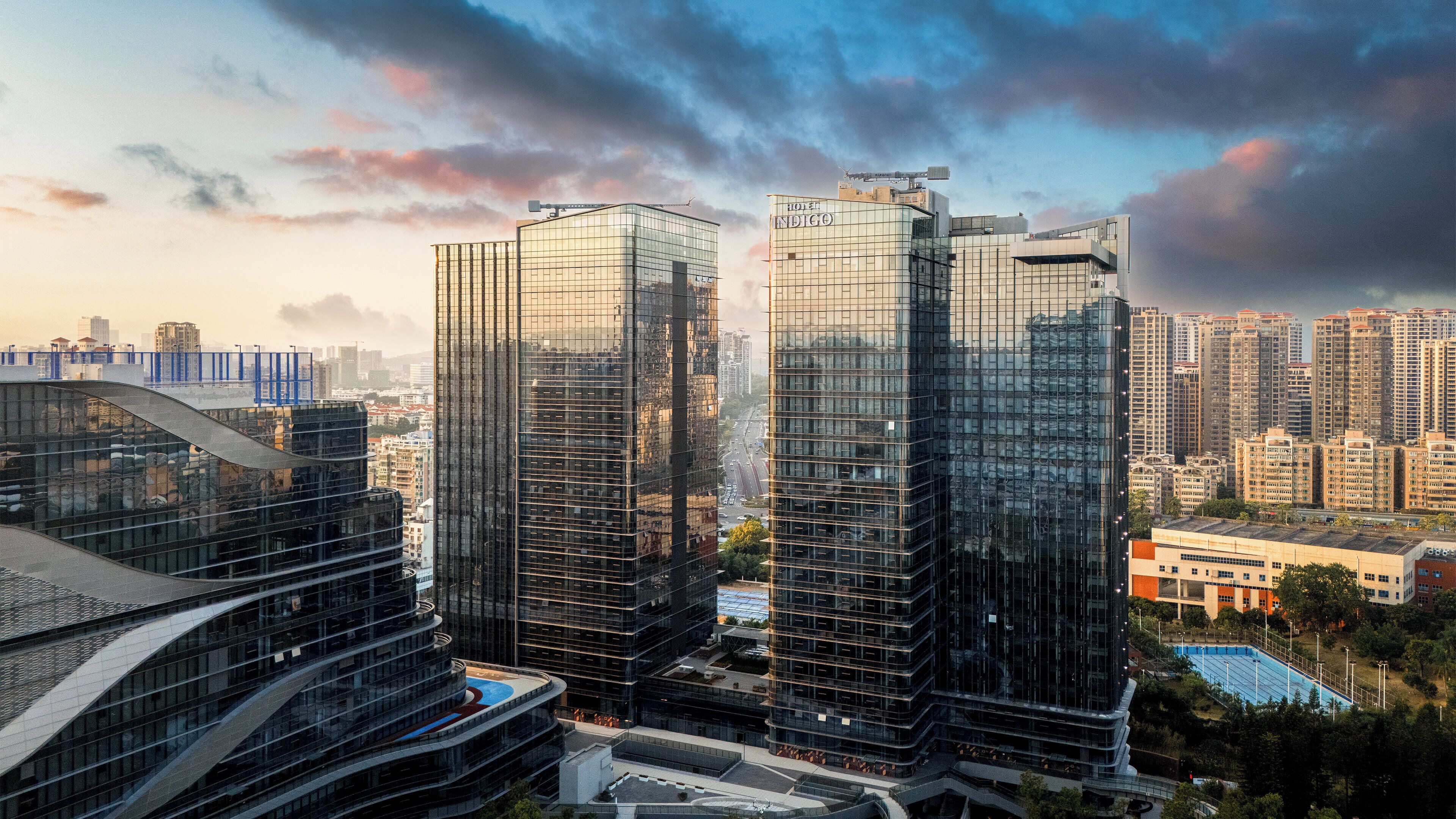 A modern high-rise hotel with a glass facade, surrounded by other high-rise buildings and a bridge in the background.