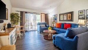 A living room with a blue sofa, a dining table with chairs, a coffee table, and a large window with curtains.