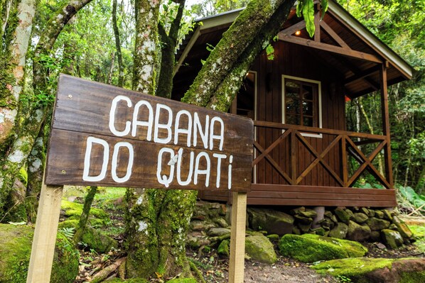 Et træskilt med teksten "CABANA DO QUATI", foran en rustik træhytte omgivet af frodigt grønt og sten.