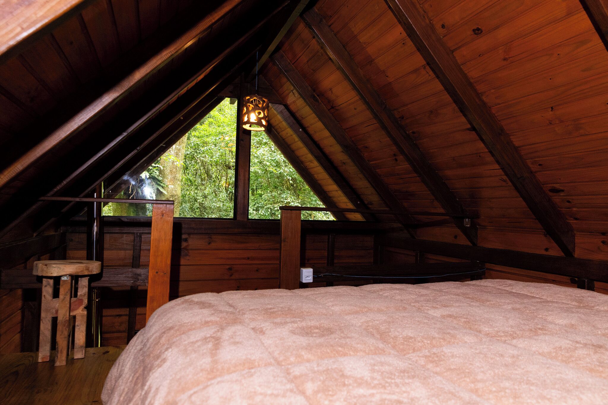 A cozy attic bedroom with a sloped ceiling, a wooden bed, a small stool, and a window with a view of greenery outside.
