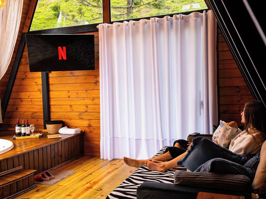 Una habitación acogedora con detalles de madera, un ventanal con cortinas, un televisor de pantalla plana en la pared, un sofá, una mesita con un control remoto y un ordenador portátil.