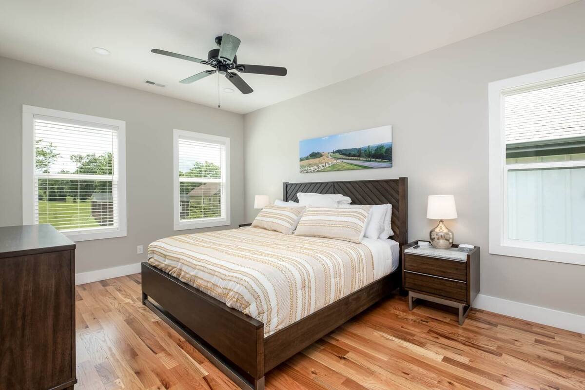 A bedroom with a bed, a nightstand, a lamp, a ceiling fan, and two windows with blinds.