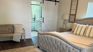 Een slaapkamer met een groot bed, een houten bankje, een witte dressoir en een schuifdeur in boerderijstijl.