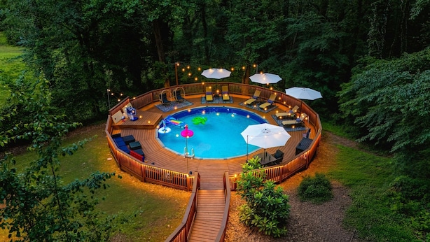 Eine runde Holzterrasse mit Pool, umgeben von Liegestühlen und Sonnenschirmen, in einem bewaldeten Gebiet.