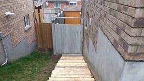 Un escalier en bois mène à une zone délimitée par une clôture, avec des murs en briques et une cour gazonnée.