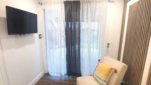 Un salon avec un téléviseur à écran plat fixé au mur, un canapé agrémenté d’un coussin à motifs, une petite table ronde avec une plante et une fenêtre ornée de rideaux légers.