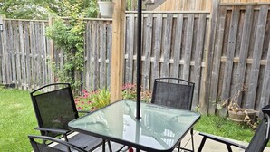 Un patio couvert avec une table en verre et quatre chaises, entouré d’une clôture en bois et de verdure.