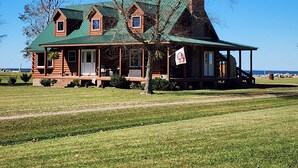 A log cabin with a green roof and a chimney, surrounded by a grassy field and trees.