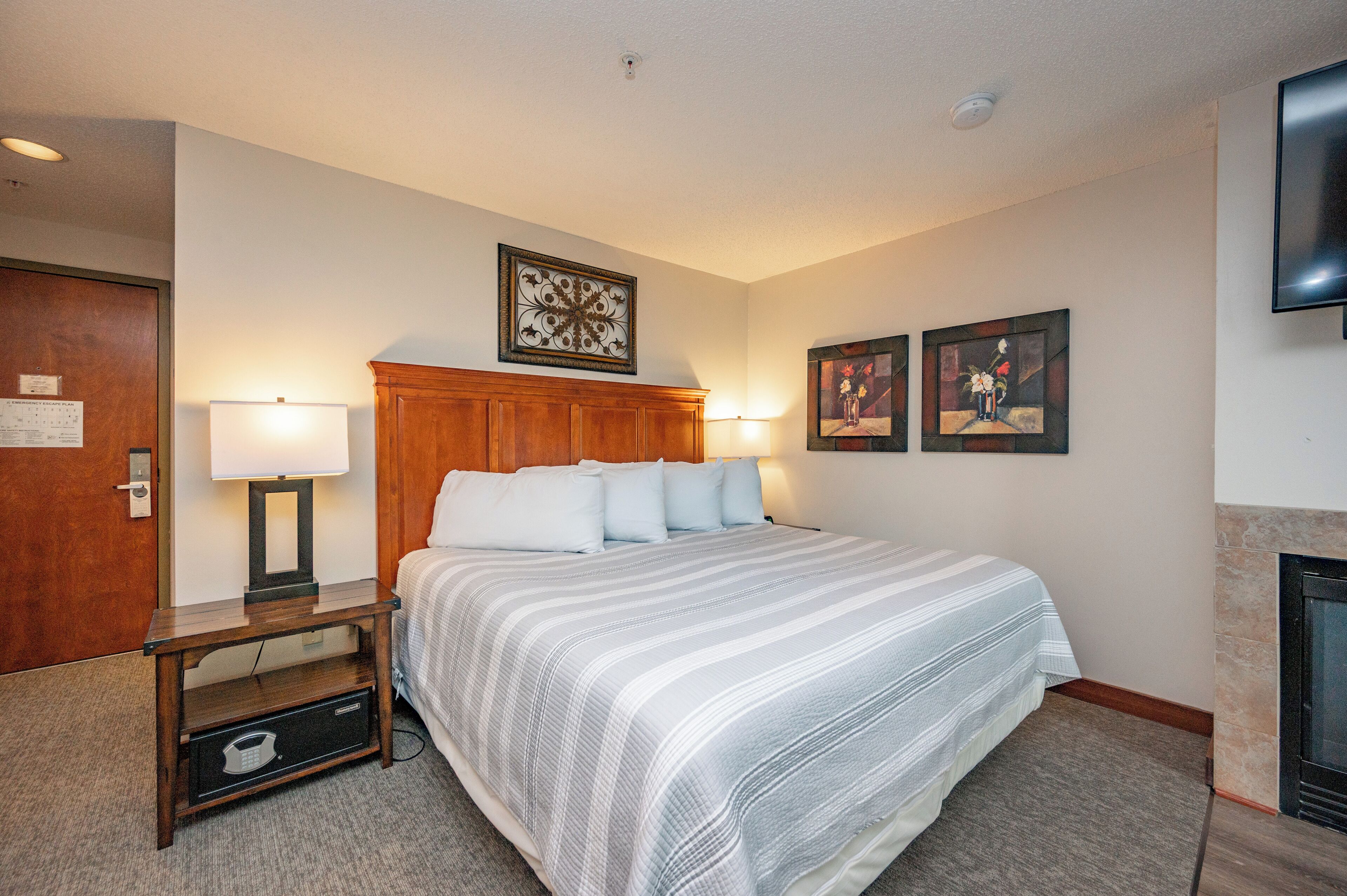 A hotel room with a large bed, a wooden nightstand with a lamp, a wall-mounted TV, and a fireplace.