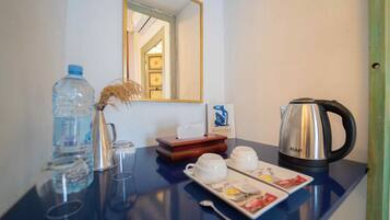 Quarto de hotel com minibar, garrafa de água, vaso com plantas secas, cafeteira e bandeja com chá e petiscos.