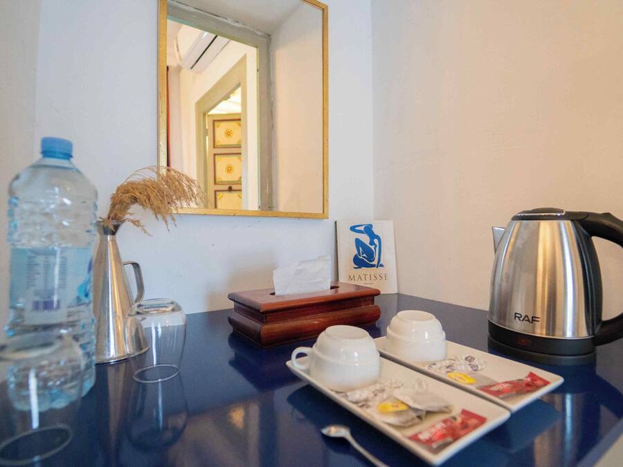Quarto de hotel com minibar, garrafa de água, vaso com plantas secas, cafeteira e bandeja com chá e petiscos.