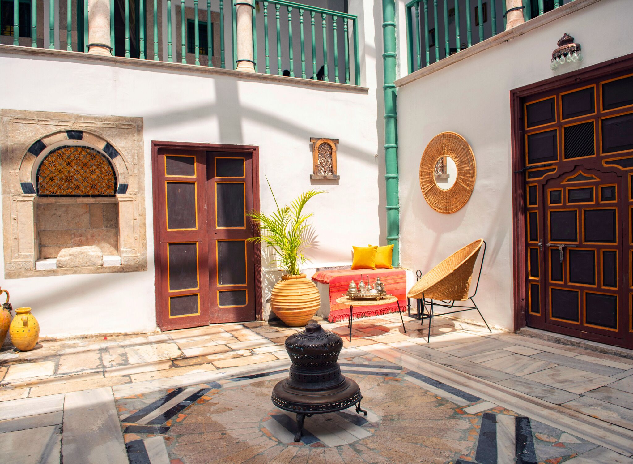 A courtyard with a tiled floor, a traditional lamp, a potted plant, a wicker chair, and a red and yellow striped rug.