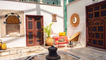 A courtyard with a tiled floor, a traditional lamp, a potted plant, a wicker chair, and a red and yellow striped rug.